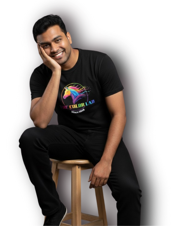 A smiling Indian male model sitting on a wooden stool in a professional photo studio, wearing a black The Color Lab t-shirt featuring a large colorful unicorn logo.